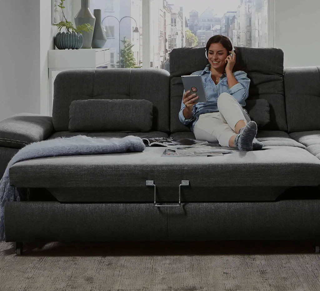 Frau entspannt auf einem grauen Sofa mit verstellbarer Rückenlehne, Tablet und Kopfhörern. Modernes Wohnzimmer mit komfortabler Couch für entspanntes Wohnen und stilvolles Ambiente.